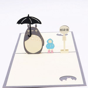 Pu&ograve; includere: Un biglietto pop-up 3D con un personaggio di Totoro grigio e bianco che tiene un ombrello nero, una bambina con un impermeabile blu e un cartello che dice "Fermata dell'autobus".