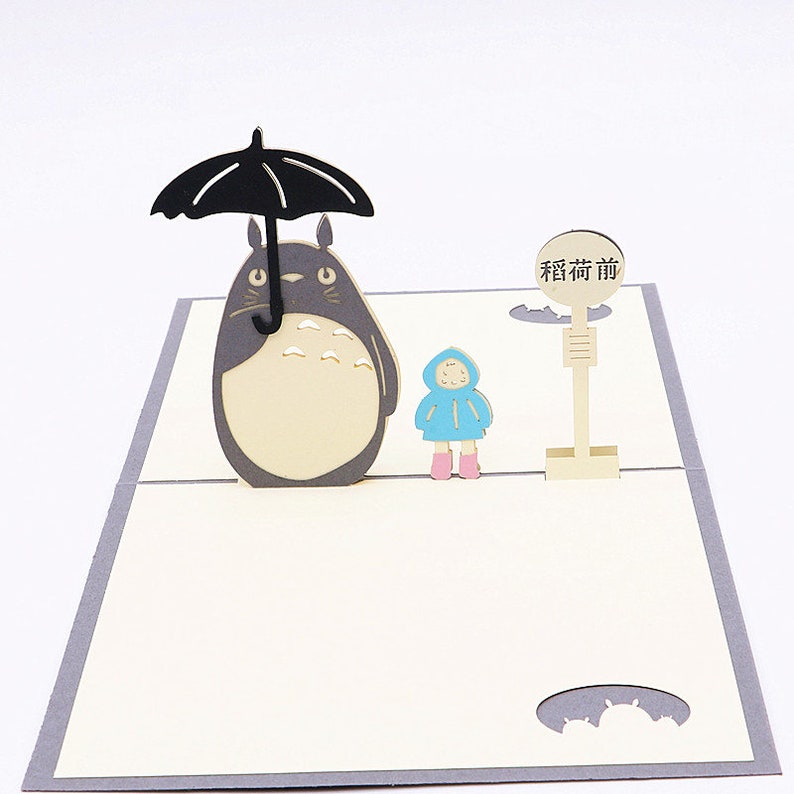 Pu&ograve; includere: Un biglietto pop-up 3D con un personaggio di Totoro grigio e bianco che tiene un ombrello nero, una bambina con un impermeabile blu e un cartello che dice "Fermata dell'autobus".