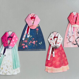 May include: Four paper crafts of traditional Korean hanbok dresses. Each dress is decorated with different floral patterns and colors, including pink, blue, red, and white.
