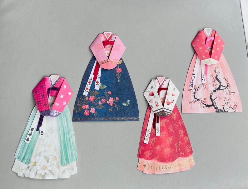 May include: Four paper crafts of traditional Korean hanbok dresses. Each dress is decorated with different floral patterns and colors, including pink, blue, red, and white.