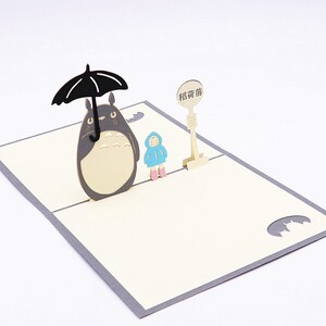 Pu&ograve; includere: Un biglietto pop-up 3D con Totoro, un personaggio del film d'animazione Il mio vicino Totoro, che tiene un ombrello sopra una piccola figura. Il biglietto ha uno sfondo grigio chiaro e un interno color crema con il testo "Guarda prima di te" in caratteri cinesi.