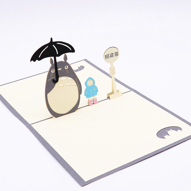 Pu&ograve; includere: Un biglietto pop-up 3D con Totoro, un personaggio del film d'animazione Il mio vicino Totoro, che tiene un ombrello sopra una piccola figura. Il biglietto ha uno sfondo grigio chiaro e un interno color crema con il testo "Guarda prima di te" in caratteri cinesi.
