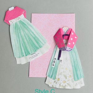 May include: Two paper cutouts of traditional Korean hanbok dresses. The dress on the left has a pink top and a green skirt. The dress on the right has a pink top with a floral pattern and a green skirt with white flowers. The text "Style C" is printed below the dresses.