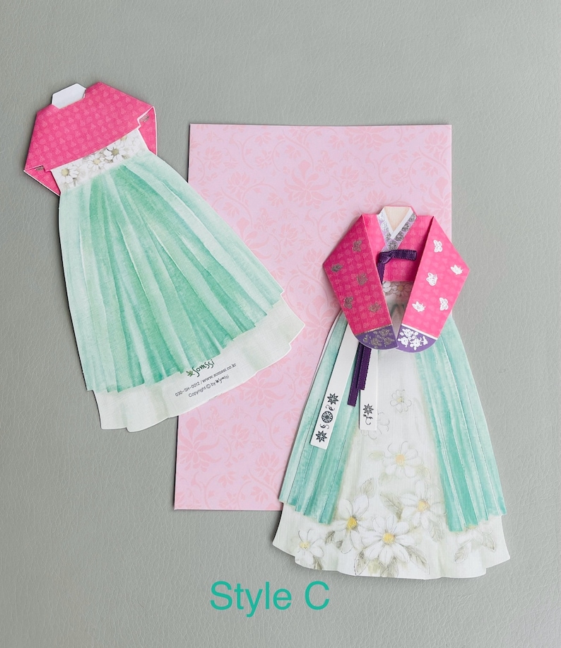 May include: Two paper cutouts of traditional Korean hanbok dresses. The dress on the left has a pink top and a green skirt. The dress on the right has a pink top with a floral pattern and a green skirt with white flowers. The text "Style C" is printed below the dresses.