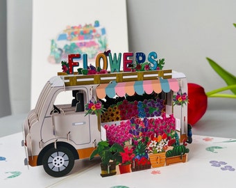 Peut inclure: Une carte pop-up blanche représentant un camion de fleurs avec une enseigne colorée qui dit "FLOWERS". Le camion est rempli de fleurs et de plantes colorées en pots. La carte est décorée d'un motif floral.