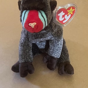May include: A gray and brown Ty Beanie Baby monkey with a red and blue face. The monkey is sitting on a brown surface. The tag says "Beanie Babies" and "Ty".