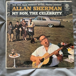 May include: Vintage vinyl record album cover for Allan Sherman's "My Son, The Celebrity." The cover features a photo of Sherman playing a guitar, with a group of people and a car in the background. The album is from Warner Bros.