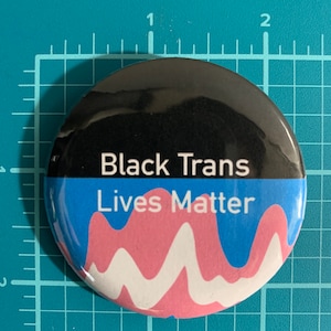 Puede incluir: Un botón redondo con una mitad superior negra y una mitad inferior azul. El texto "Black Trans" está en blanco en la sección negra, y "Lives Matter" está en blanco en la sección azul. Un diseño ondulado rosa y blanco está en la sección azul.