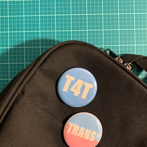 May include: Two round buttons with text on a black surface. One button is light blue with the white text "T4T". The other button has a pink and light blue background with the white text "TRANS*".