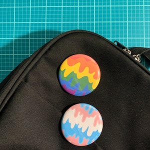 May include: Two round buttons with colorful designs. One button features a rainbow dripping design, and the other has a pink, white, and blue dripping design. The buttons are placed on a black bag.