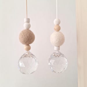 Könnte beinhalten: Zwei hängende Ornamente mit facettierten, klaren Kristallkugeln an weißen Schnüren. Jedes Ornament besteht aus einer Abfolge von Perlen: einem kleinen weißen Zylinder, einer strukturierten weißen Kugel, einer großen Filzkugel in Beige oder Creme.