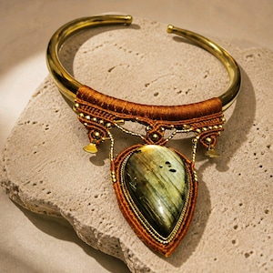 May include: A gold-toned metal torque necklace featuring a large, pear-shaped, iridescent grey and green gemstone pendant. The pendant is framed by intricate, dark orange thread wrapping and small gold-toned metal beads and accents.