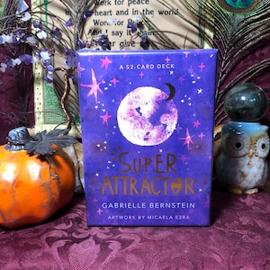 May include: A 52-card deck titled "Super Attractor" by Gabrielle Bernstein. The box is purple with a large white moon, stars, and gold lettering. The artwork is by Micaela Ezra. The deck is displayed with a decorative pumpkin and owl figurine.