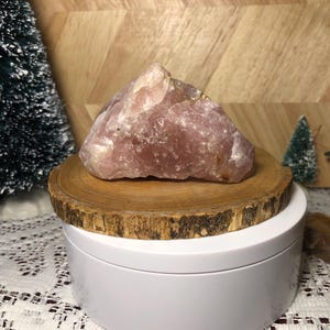 May include: A large, rough-cut rose quartz crystal, displaying a soft pink hue, rests on a natural wood slice. The crystal is positioned on a white, rotating display stand, with a wooden background and small decorative trees.