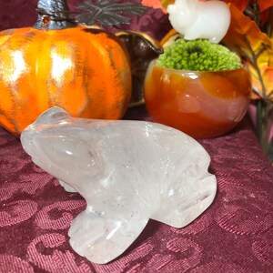 May include: A clear quartz frog figurine on a burgundy fabric background. The frog is carved from a single piece of quartz and has a smooth, polished finish.