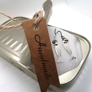 Può includere: Un paio di orecchini a forma di pesce argentati con dettagli dorati, confezionati in una scatola di latta argentata con un'etichetta marrone che dice "Handmade".