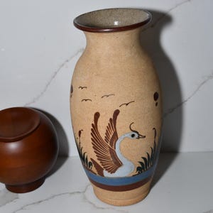 May include: Tan ceramic vase with a swan design. The vase features a brown rim and base, with a blue stripe and brown and white swan illustration. The vase also has a wooden bowl next to it.