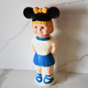May include: Vintage plastic doll wearing a black Mickey Mouse ears hat with a yellow bow. The doll has blonde hair, a white shirt with a blue collar, a blue skirt, and blue shoes. The doll is standing upright.