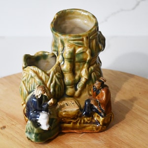 May include: A ceramic vase with a bamboo-like design in shades of green and brown. Two figures are seated, appearing to play a board game. The vase has two openings and is placed on a wooden surface.