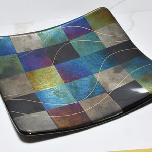 May include: Square decorative glass plate with a checkered pattern in shades of blue, green, and gray. The plate has a black border and gold-colored wavy lines. A unique piece for serving or display.