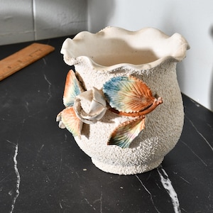 May include: A white ceramic planter with a textured surface and a ruffled rim. The planter features a 3D floral design with orange, blue, and white accents.