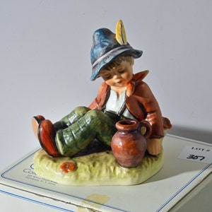 May include: A porcelain figurine of a boy wearing a blue hat with a feather, a red jacket, and green pants. He is sitting on a grassy base with a brown jug beside him. The figurine is part of a collection and is in a box with the lot number 307.