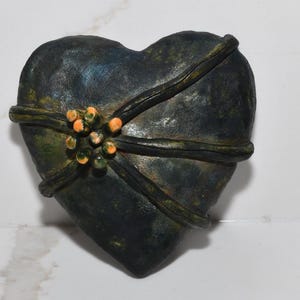 May include: A dark green heart-shaped decorative object with a textured surface. The heart is wrapped with a darker band and adorned with a cluster of orange and green beads. The object is set against a white background.