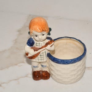 May include: A ceramic planter featuring a young girl figurine with orange hair and a blue dress, playing a small instrument. The planter has a basket-weave design and a blue rim. The figurine is approximately 4 inches tall.