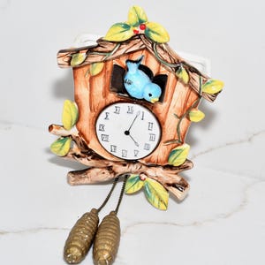 May include: A ceramic cuckoo clock in a rustic design. The clock features a brown wooden house with a blue bird, a white clock face, and decorative leaves. The clock has two pinecone-shaped weights hanging below.
