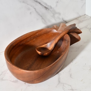 May include: A wooden serving bowl and spoon set. The bowl is oval-shaped with a textured exterior and a spoon rests inside. The set is made of a dark brown wood, and the spoon is curved to fit the bowl. The bowl has a pineapple-like design.