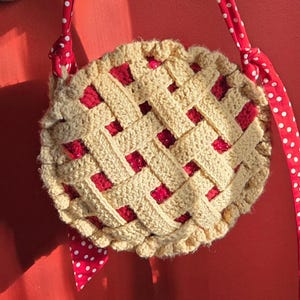 Könnte beinhalten: Runde, gehäkelte Tasche, die einem Kuchen ähnelt, mit einem beigefarbenen Gittermuster und roter Füllung. Sie hat einen rot-weiß gepunkteten Riemen. Die Tasche hat einen Durchmesser von etwa 20 cm.
