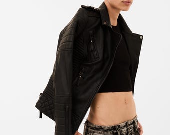 May include: A black leather motorcycle jacket featuring quilted paneling on the shoulders and sleeves, a notched lapel, silver-tone metal zippers, and a front zip closure, worn over a black cropped top and acid-wash gray denim jeans.