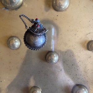 May include: A silver and red ornate bottle with a chain attached. The bottle is round and has intricate designs. The bottle is sitting on a metal surface with a pattern of round indentations.