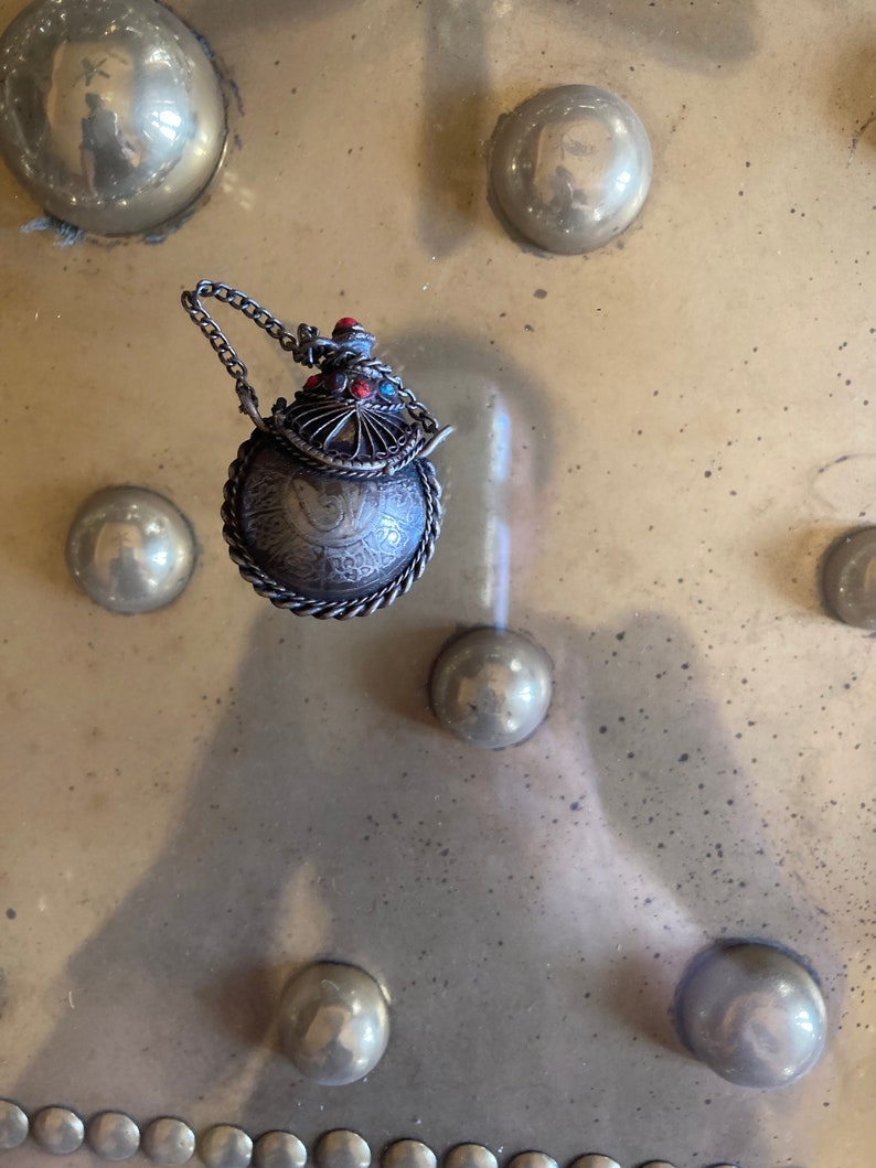 May include: A silver and red ornate bottle with a chain attached. The bottle is round and has intricate designs. The bottle is sitting on a metal surface with a pattern of round indentations.