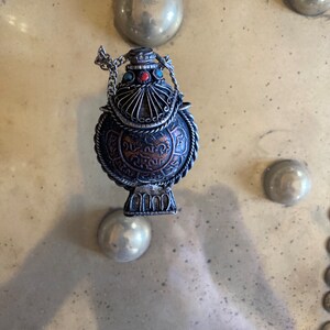 May include: A silver and copper colored metal bottle with a chain and intricate designs. The bottle has a round body and a small opening at the top. The bottle is decorated with turquoise and red stones.