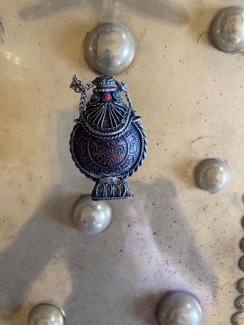 May include: A silver and copper colored metal bottle with a chain and intricate designs. The bottle has a round body and a small opening at the top. The bottle is decorated with turquoise and red stones.