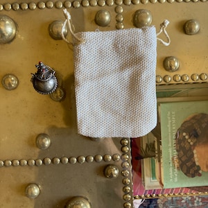 May include: A small, ornate, silver-colored, jeweled, bird-shaped pendant in a small, off-white, drawstring pouch.