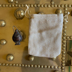 May include: A small, round, silver and copper colored metal bottle with a red stone on the top. The bottle is hanging from a silver chain and is sitting on a gold surface. The bottle is in a small, white, fabric bag with a drawstring.
