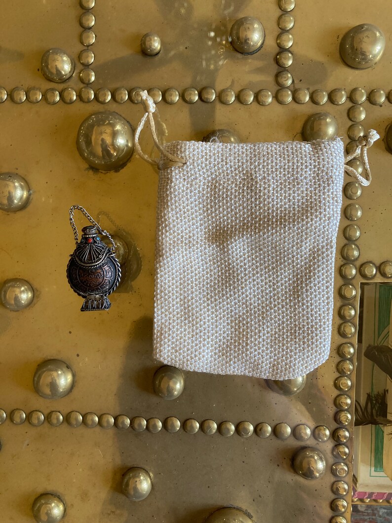 May include: A small, round, silver and copper colored metal bottle with a red stone on the top. The bottle is hanging from a silver chain and is sitting on a gold surface. The bottle is in a small, white, fabric bag with a drawstring.