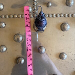 May include: A silver and copper colored metal bottle with intricate designs and a small chain. The bottle is about 2 inches tall and is sitting on a gold surface with a pattern of small circles.