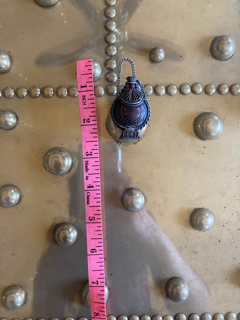 May include: A silver and copper colored metal bottle with intricate designs and a small chain. The bottle is about 2 inches tall and is sitting on a gold surface with a pattern of small circles.