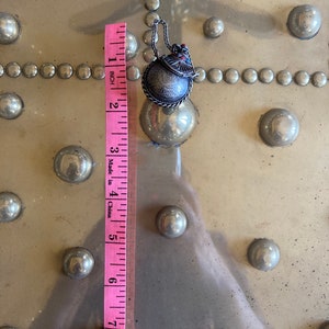 May include: A silver charm with a round, engraved design and a red and blue enamel detail. The charm is attached to a silver chain and measures approximately 1 inch in diameter.