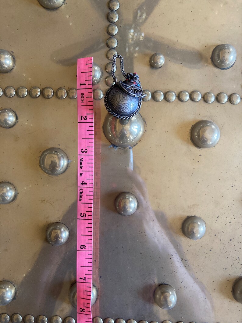 May include: A silver charm with a round, engraved design and a red and blue enamel detail. The charm is attached to a silver chain and measures approximately 1 inch in diameter.