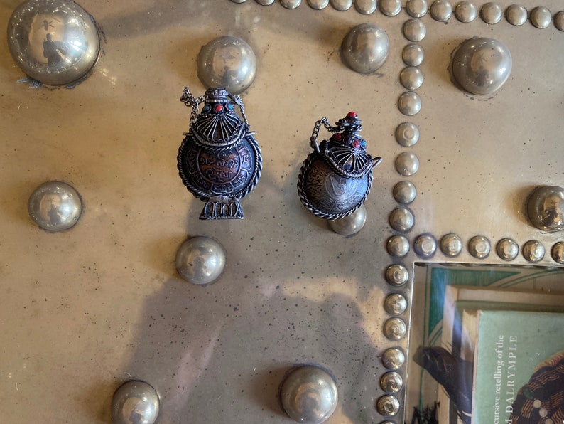 May include: Two silver and copper colored decorative teapots with red and blue accents. The teapots are on a gold background with a pattern of round circles.