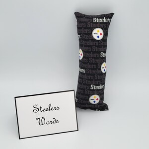 May include: A black and gray fabric pillow with the word "Steelers" repeated in a pattern. The pillow has a Steelers logo with a yellow star and a red and blue stripe. The pillow is sitting on a white surface with a white card that says "Steelers Words" in black cursive writing.
