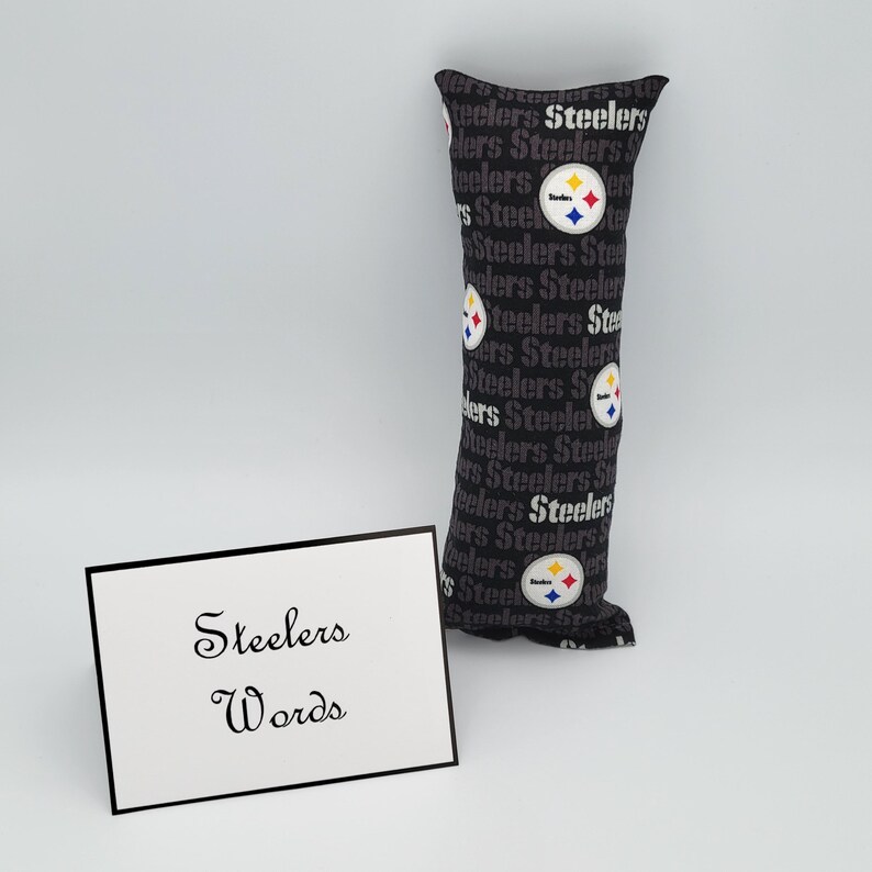 May include: A black and gray fabric pillow with the word "Steelers" repeated in a pattern. The pillow has a Steelers logo with a yellow star and a red and blue stripe. The pillow is sitting on a white surface with a white card that says "Steelers Words" in black cursive writing.