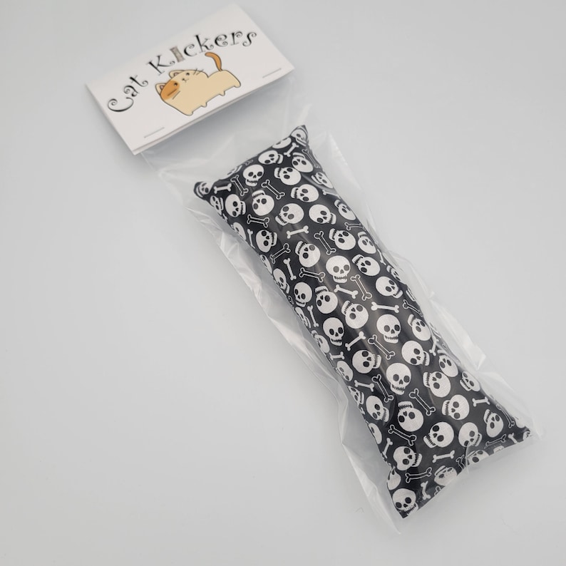 May include: A black cat toy with a white skull and crossbones pattern. The toy is packaged in a clear plastic bag with a white label that says "Cat Kickers".