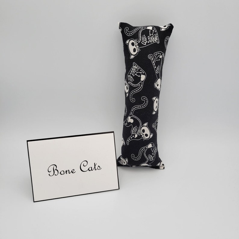 May include: Black cat-shaped catnip toy with a white skeleton design. The toy is made of fabric and has a rectangular shape. The text "Bone Cats" is printed on a white card.