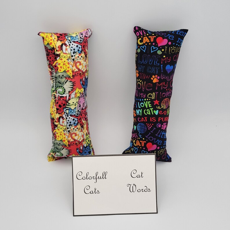 May include: Two cat-themed fabric toys. One toy is black with colorful text that says "I love my cat" and other cat-related phrases. The other toy is red with a colorful pattern of cats. Both toys are long and rectangular.