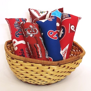 May include: A wicker basket filled with five colorful fabric pillows. The pillows have a baseball theme with red, white, and blue colors. The pillows have text on them, including "Phillies" and "Philadelphia".